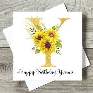 May include: A white birthday card featuring a gold letter "Y" adorned with sunflowers and foliage. The card displays the message "Happy Birthday Yvonne" in black script. The card is presented with a white envelope.