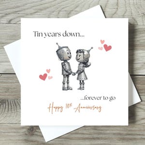 May include: A white greeting card with a drawing of two robot figures holding hands, surrounded by pink hearts. The card reads "Tin years down...forever to go" and "Happy 10th Anniversary" in a handwritten style font.