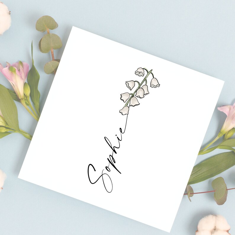 May include: A white card with the name "Sophie" written in black cursive script. A delicate illustration of a white lily of the valley flower with green stems is positioned above the name.
