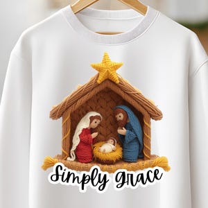 Puede incluir: Sudadera blanca con un pesebre tejido. María está vestida de rojo, José de azul y el niño Jesús en un pesebre. Una estrella amarilla está encima del establo marrón. El texto "Simply grace" está debajo.