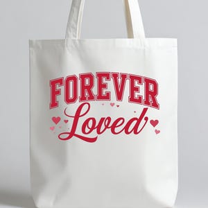 May include: A white tote bag with the words "FOREVER Loved" in red lettering. The word "FOREVER" is in a bold, blocky font, while "Loved" is in a cursive script. Small pink hearts are scattered around the text.