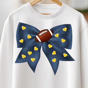 May include: White sweatshirt featuring a large navy blue bow adorned with yellow hearts. A brown football is centered on the bow. The sweatshirt is hanging on a wooden hanger.
