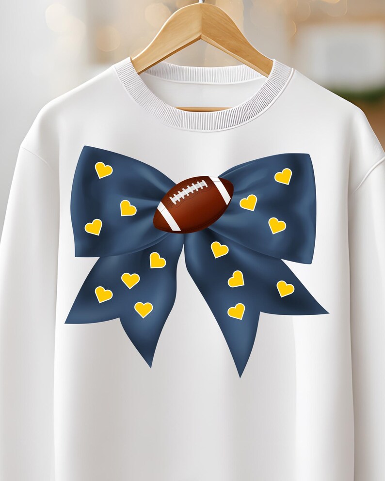 May include: White sweatshirt featuring a large navy blue bow adorned with yellow hearts. A brown football is centered on the bow. The sweatshirt is hanging on a wooden hanger.