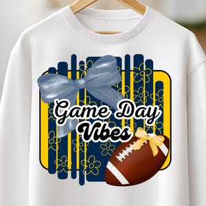 May include: White sweatshirt with a graphic design featuring a blue bow, a football with a yellow bow, and the text "Game Day Vibes" in a black font. The design includes yellow and blue floral elements.