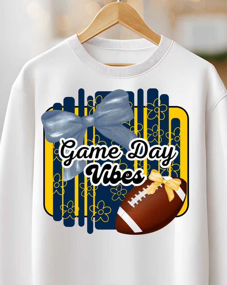 May include: White sweatshirt with a graphic design featuring a blue bow, a football with a yellow bow, and the text "Game Day Vibes" in a black font. The design includes yellow and blue floral elements.