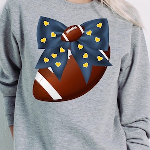 May include: A gray sweatshirt featuring a football design. A navy blue bow with yellow hearts is placed over a brown football. The football has white stripes and laces. The design is centered on the chest of the sweatshirt.