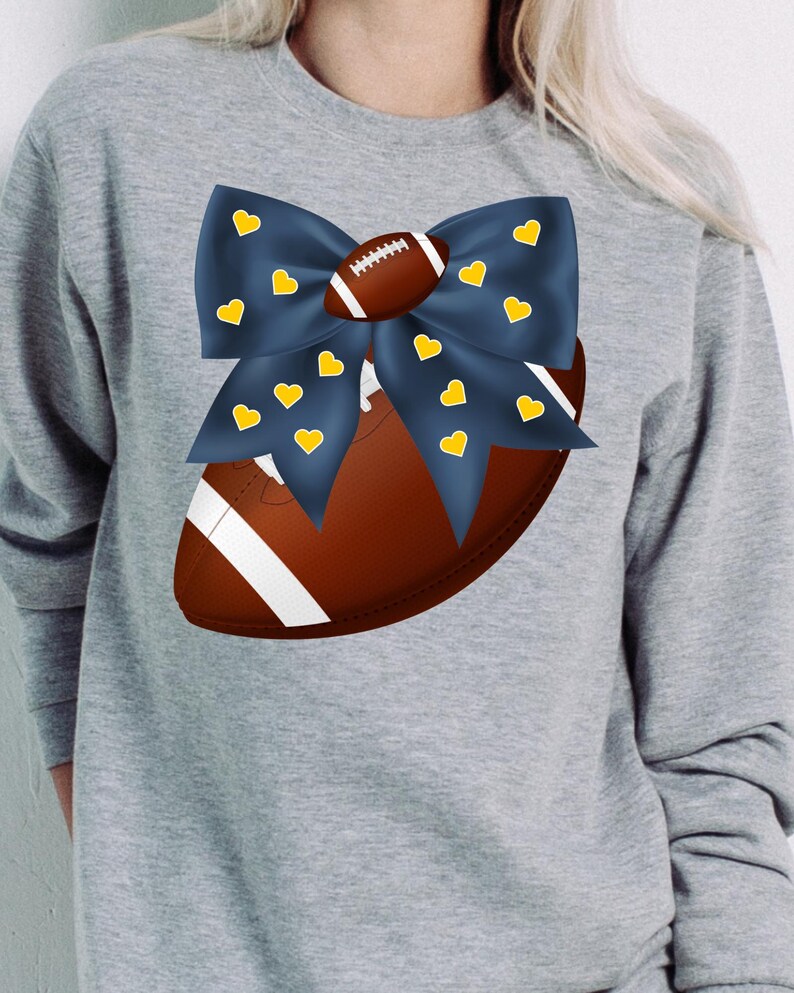 May include: A gray sweatshirt featuring a football design. A navy blue bow with yellow hearts is placed over a brown football. The football has white stripes and laces. The design is centered on the chest of the sweatshirt.