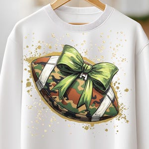 May include: White sweatshirt featuring a football design with a camouflage pattern and a green bow. The football is outlined with gold glitter and has white stripes. The sweatshirt is hanging on a wooden hanger.
