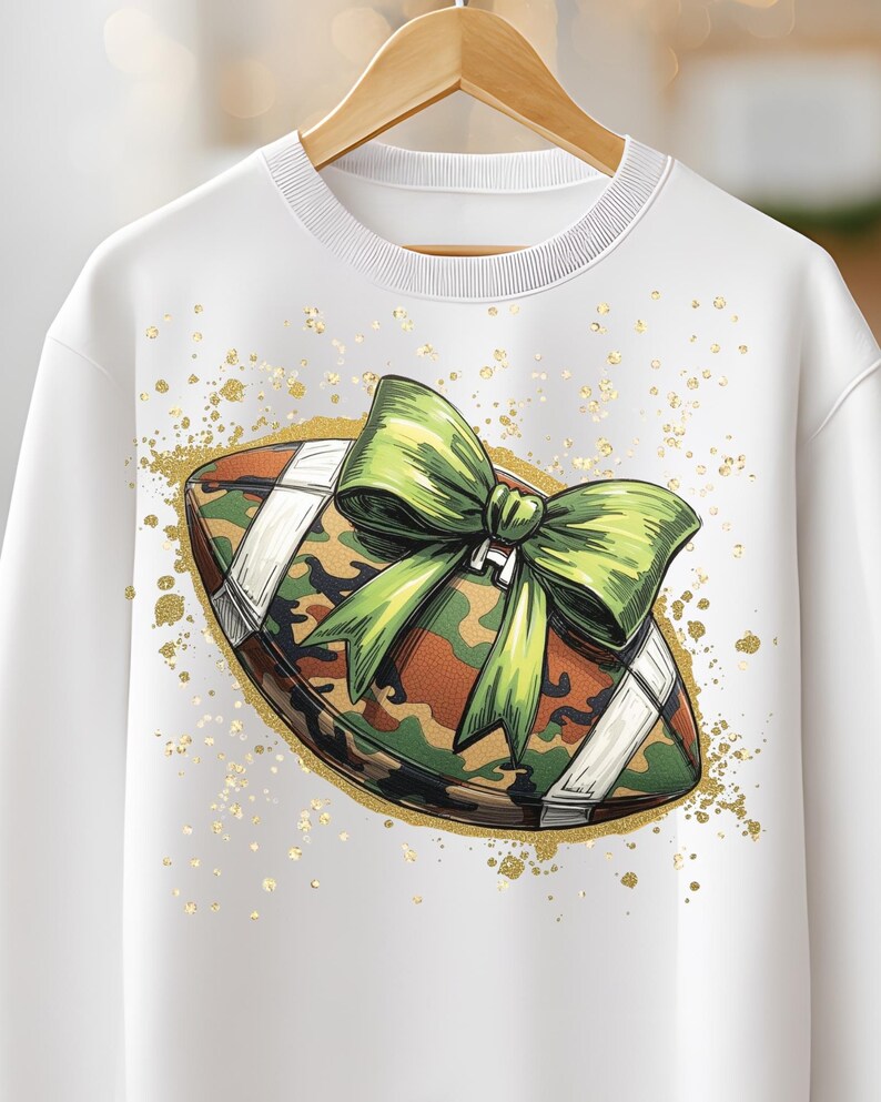 May include: White sweatshirt featuring a football design with a camouflage pattern and a green bow. The football is outlined with gold glitter and has white stripes. The sweatshirt is hanging on a wooden hanger.