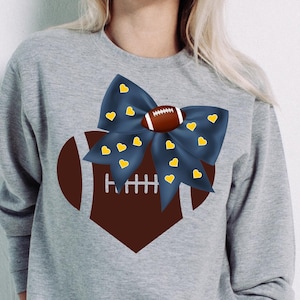 May include: A gray sweatshirt featuring a football-themed design. A dark blue bow with yellow hearts is centered above a brown football shape. The football has white laces. The sweatshirt is a casual, sporty style.