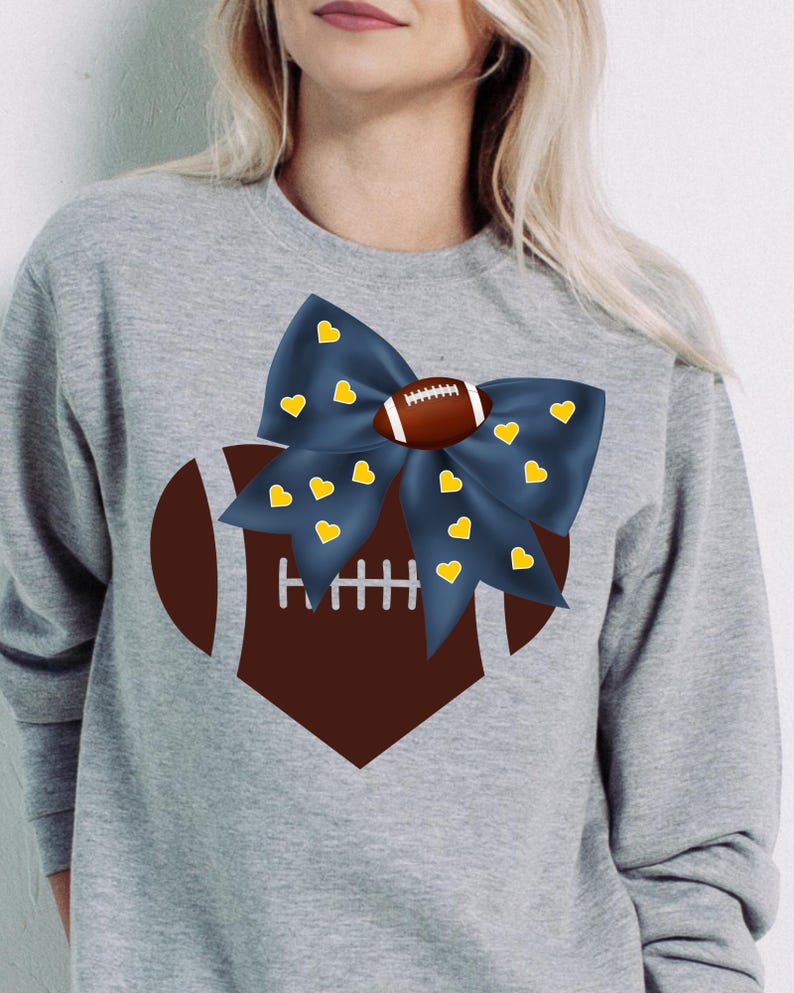 May include: A gray sweatshirt featuring a football-themed design. A dark blue bow with yellow hearts is centered above a brown football shape. The football has white laces. The sweatshirt is a casual, sporty style.