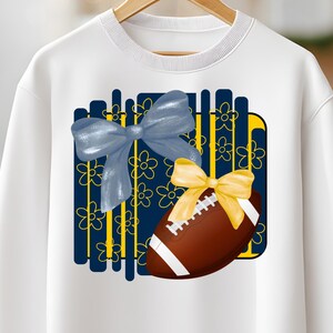 May include: White sweatshirt featuring a graphic of a brown football with white stripes and a yellow bow. A blue bow and floral design are also present. The sweatshirt is hanging on a wooden hanger.