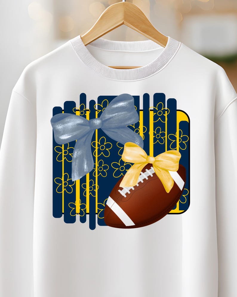 May include: White sweatshirt featuring a graphic of a brown football with white stripes and a yellow bow. A blue bow and floral design are also present. The sweatshirt is hanging on a wooden hanger.
