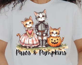 May include: White t-shirt featuring a whimsical illustration of three cartoon cats. One cat is dressed in a princess gown, another in knight armor, and the third is in a jack-o'-lantern. The text "Paws & Pumpkins" is below the illustration.