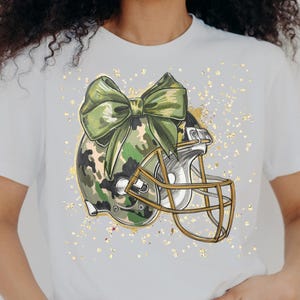 May include: White t-shirt featuring a football helmet with a camouflage pattern and a large green bow. The helmet has a gold-colored face mask. Gold glitter-like accents are scattered around the design. The shirt is a casual, sporty style.
