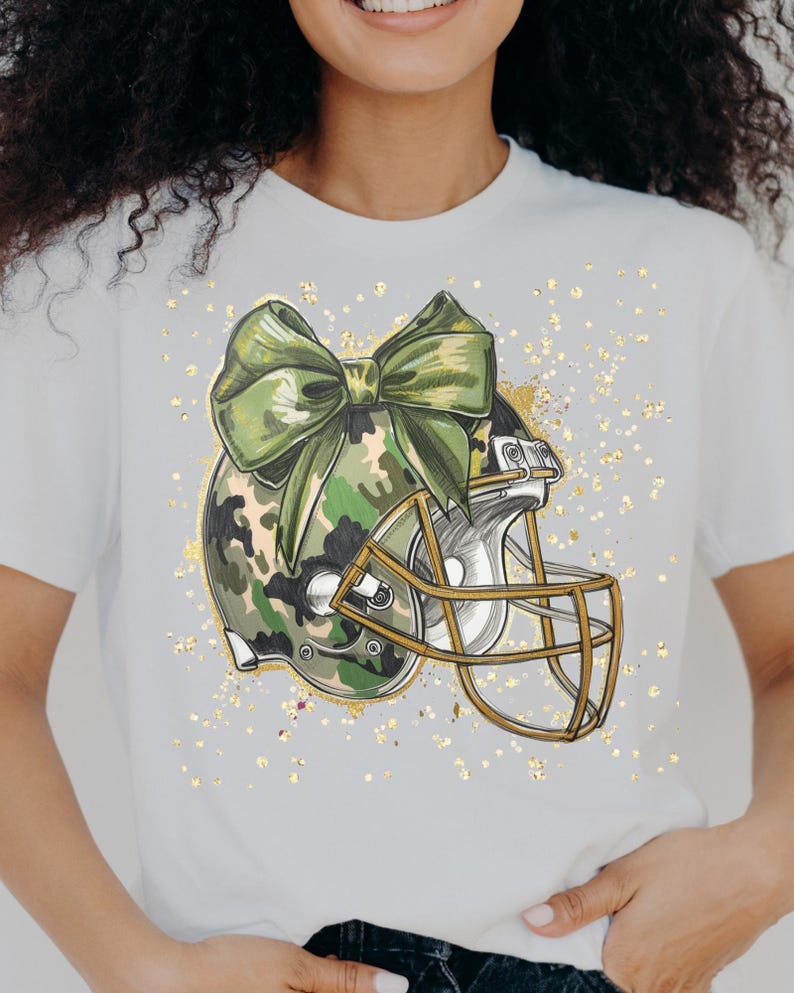 May include: White t-shirt featuring a football helmet with a camouflage pattern and a large green bow. The helmet has a gold-colored face mask. Gold glitter-like accents are scattered around the design. The shirt is a casual, sporty style.