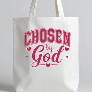 Puede incluir: Un bolso tote blanco con el texto rosa "CHOSEN by God". La palabra "CHOSEN" está en una fuente de estilo universitario, y "God" está en cursiva. Pequeños corazones rosas salpican el texto.