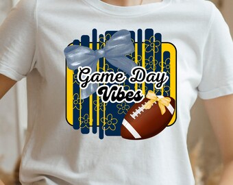 May include: White t-shirt featuring a graphic with a blue bow, a football, and the text "Game Day Vibes." The design includes a navy blue and yellow background with floral accents, perfect for football fans.