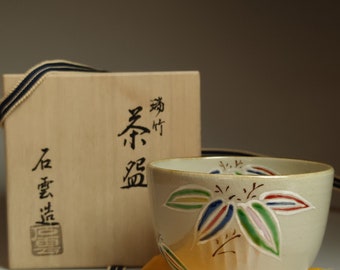 May include: A light grey ceramic teacup with a floral design in shades of green, blue, red, and pink. The cup is sitting on a yellow cloth next to a wooden box with Japanese characters.