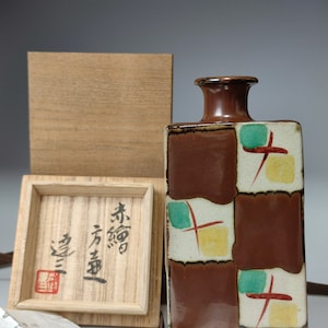 Pode incluir: Uma garrafa retangular de cerâmica marrom com gargalo marrom escuro e um padrão decorativo de quadrados marrons, brancos, verdes, amarelos e vermelhos. Uma caixa de madeira e uma caixa de madeira menor com texto japonês estão ao lado da garrafa.