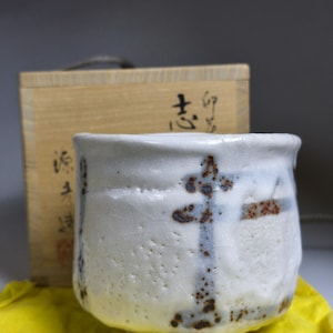 May include: A white ceramic tea cup with a textured surface and a cross-like design in blue and brown. The cup sits on a yellow cloth, with a wooden box behind it. Japanese characters are visible on the box.