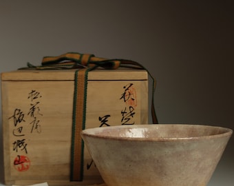 以下が含まれることがあります： わずかに不揃いな縁と粗い質感の底を持つ、薄い茶色の陶器の茶碗。茶碗は、緑と茶色のリボンと日本語の文字が書かれた木箱の横に、黄色の布の上に置かれている。