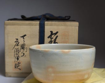 May include: A cream-glazed ceramic tea bowl with orange and pink hues, resting on a yellow cloth. A wooden box with Japanese characters and a blue ribbon is in the background. A traditional tea ceremony item.