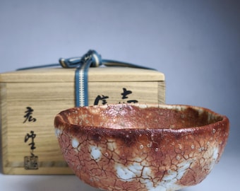 Puede incluir: Un cuenco de té de cerámica marrón y blanco con un esmalte craquelado. El cuenco está sentado sobre una caja de madera con caracteres japoneses en el lateral.