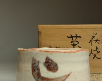 May include: A white ceramic teacup with a brown and blue design. The cup is sitting on a wooden box with Japanese characters that read "茶碗".