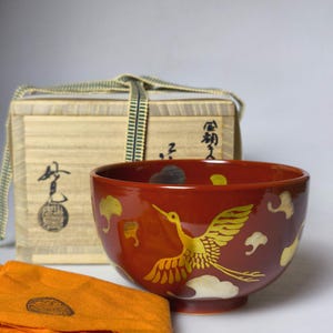 Puede incluir: Un cuenco de cerámica roja con un diseño de grulla dorada y acentos de nubes. El cuenco forma parte de un conjunto que incluye una caja de madera y un paño naranja. La grulla está en vuelo.