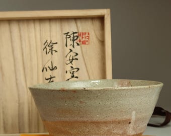 May include: A light brown ceramic tea bowl with a white glaze and a rough texture. The bowl is sitting on a yellow cloth and next to a wooden box with Japanese characters.