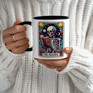 May include: White ceramic mug with a black rim and handle. The mug features a colourful illustration of a skeleton reading a book with the text "THE READER" below the image.