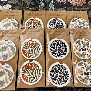 May include: Twelve round car coasters with various floral and butterfly designs. The coasters are packaged in clear plastic and labeled "Car Coasters Handmade with Love".