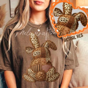 Può includere: T-shirt marrone con un design a coniglio patchwork. Il coniglio presenta sezioni con stampa leopardata, a righe e in tessuto marrone tinta unita. Il coniglio ha lunghe orecchie ed è centrato sulla maglietta. Il testo "Thelacrafts" è visibile.