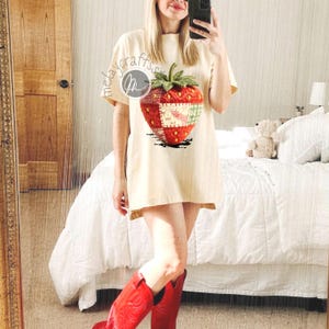 Puede incluir: Vestido camiseta color crema con un gran gráfico de fresa. El diseño de la fresa incorpora elementos de patchwork en rojo, verde y blanco. La persona lleva botas de vaquero rojas.