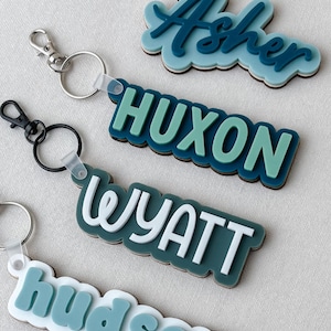 May include: Four custom keychains featuring layered acrylic name plates in shades of blue, teal, and white. Each keychain has a metal lobster clasp and ring, displaying the names Asher, HUXON, WYATT, and hudson in various fonts and color combinations.