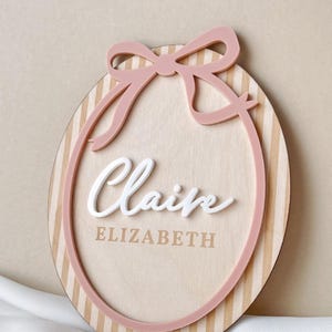 Puede incluir: Letrero ovalado de madera con lazo y borde rosas. El letrero tiene un fondo de vetas de madera clara con rayas verticales. El nombre "Claire" est&aacute; en escritura blanca, y "ELIZABETH" est&aacute; impreso debajo.