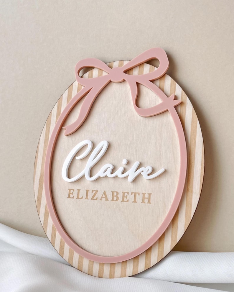 Puede incluir: Letrero ovalado de madera con lazo y borde rosas. El letrero tiene un fondo de vetas de madera clara con rayas verticales. El nombre "Claire" est&aacute; en escritura blanca, y "ELIZABETH" est&aacute; impreso debajo.