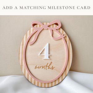 Puede incluir: Una tarjeta de hito de madera para el cuarto mes de un beb&eacute;. La tarjeta redonda tiene un lazo y un marco rosas, con el n&uacute;mero 4 en blanco y la palabra "months" en escritura. El fondo tiene rayas verticales.