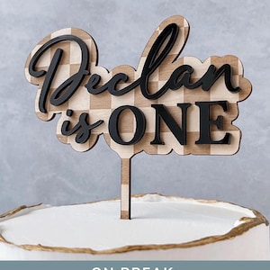 May include: A wooden cake topper featuring a light brown and beige checkered pattern background with the black text "Declan is ONE" layered on top. The topper has a single vertical stake and is positioned on top of a white frosted cake.