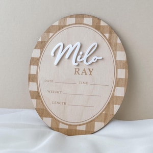 K&ouml;nnte beinhalten: Ein ovales Geburtsank&uuml;ndigungsschild aus Holz mit einem beigefarbenen und wei&szlig;en Karomuster am Rand. Der Name Milo ist in erhabener wei&szlig;er Schrift dargestellt, darunter steht Ray. Es enth&auml;lt Zeilen f&uuml;r Date, Time, Weight und Length.
