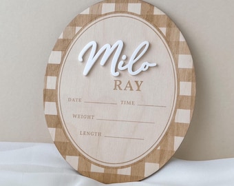 May include: An oval wooden birth announcement sign featuring a tan and white checkered border. The name Milo is in raised white script, with Ray printed below. It includes lines for Date, Time, Weight, and Length for recording newborn details.