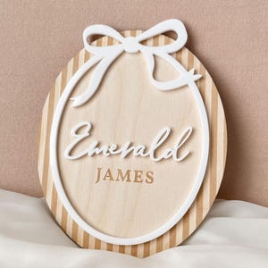 Puede incluir: Letrero ovalado de madera con lazo y borde blancos. El letrero tiene un fondo de vetas de madera clara con rayas verticales. El nombre "Emerald" est&aacute; escrito en cursiva, con "JAMES" debajo en fuente de bloque.