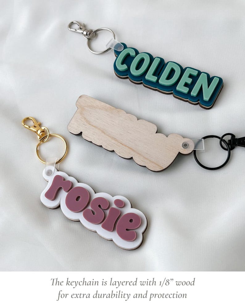 May include: Three custom keychains featuring layered designs. One displays the name COLDEN in light green on a dark teal base with a silver clip. Another is a plain wooden cutout. The third shows the name rosie in mauve on a white base with a gold clip.