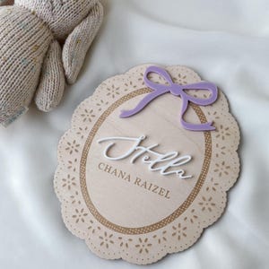 May include: An oval wooden plaque with a scalloped edge featuring a laser-cut floral pattern, a purple acrylic bow, and the white 3D script text "Hello" above the engraved name "CHANA RAIZEL". A beige knit plush toy is positioned nearby.