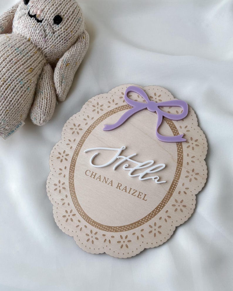 May include: An oval wooden plaque with a scalloped edge featuring a laser-cut floral pattern, a purple acrylic bow, and the white 3D script text "Hello" above the engraved name "CHANA RAIZEL". A beige knit plush toy is positioned nearby.