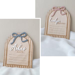 May include: Two arched wooden birth announcement signs with vertical stripes. One features a blue bow and the name Asher Alin, the other a pink bow and the name Lendyn Rhodes. Both include white 3D lettering and printed lines for date, time, weight, and length.