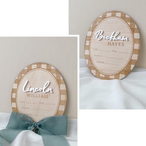 K&ouml;nnte beinhalten: Zwei ovale Geburtsank&uuml;ndigungsschilder aus Holz mit einem beigefarbenen und wei&szlig;en Karomuster am Rand. Jedes Schild zeigt einen Namen in erhabener wei&szlig;er Schrift, mit gedruckten Linien f&uuml;r Datum, Uhrzeit, Gewicht und L&auml;nge. Ein Schild hat eine salbeigr&uuml;ne Schleife.