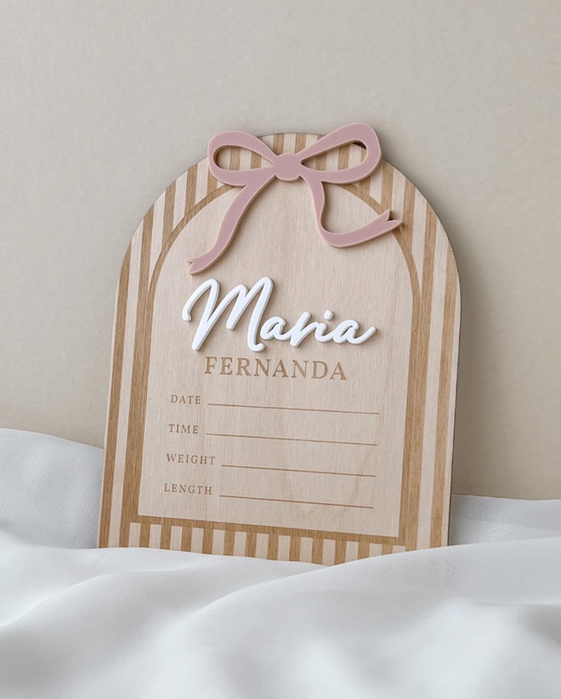 May include: An arched wooden birth announcement sign featuring a light pink acrylic bow at the top. The sign displays the name Maria in white script, the name FERNANDA in brown, and fields for DATE, TIME, WEIGHT, and LENGTH with horizontal lines.