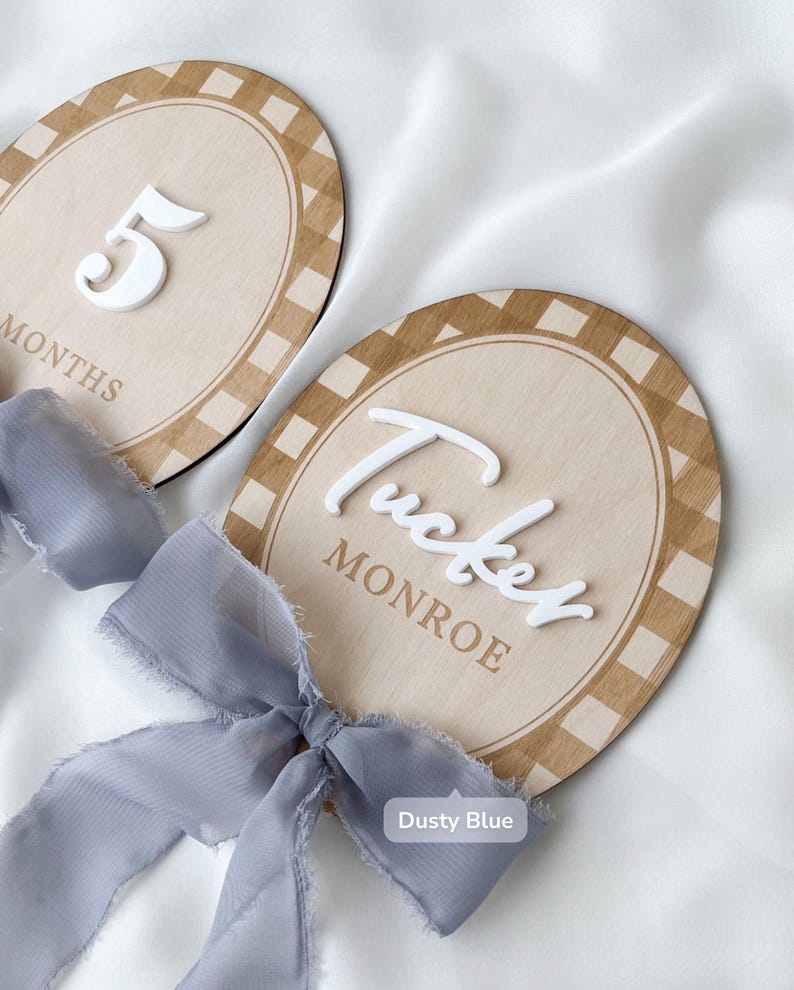 K&ouml;nnte beinhalten: Zwei ovale Holzschilder mit einem beigefarbenen und wei&szlig;en Karorand. Ein Schild zeigt die wei&szlig;e 3D-Zahl 5 und das Wort MONTHS. Das andere Schild tr&auml;gt den wei&szlig;en 3D-Namen Tucker und das Wort MONROE, beide verziert mit staubblauen Schleifen.